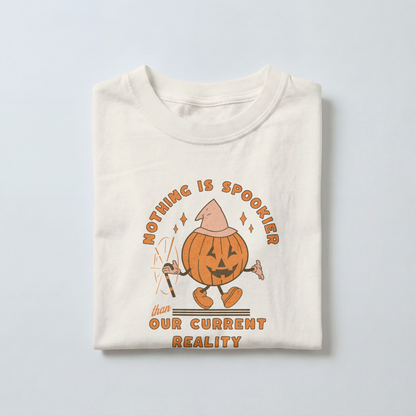 Folded ivory Comfort Colors tee with a distressed orange pumpkin wearing a peach witch hat and holding a wand. Text reads “Nothing is spookier than our current reality.”