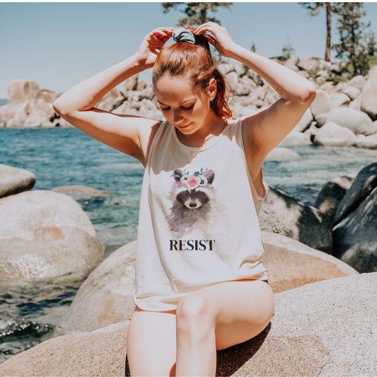 Woman sitting on rocks by a body of water wearing a tank top with a raccoon graphic and 'RESIST' text.
